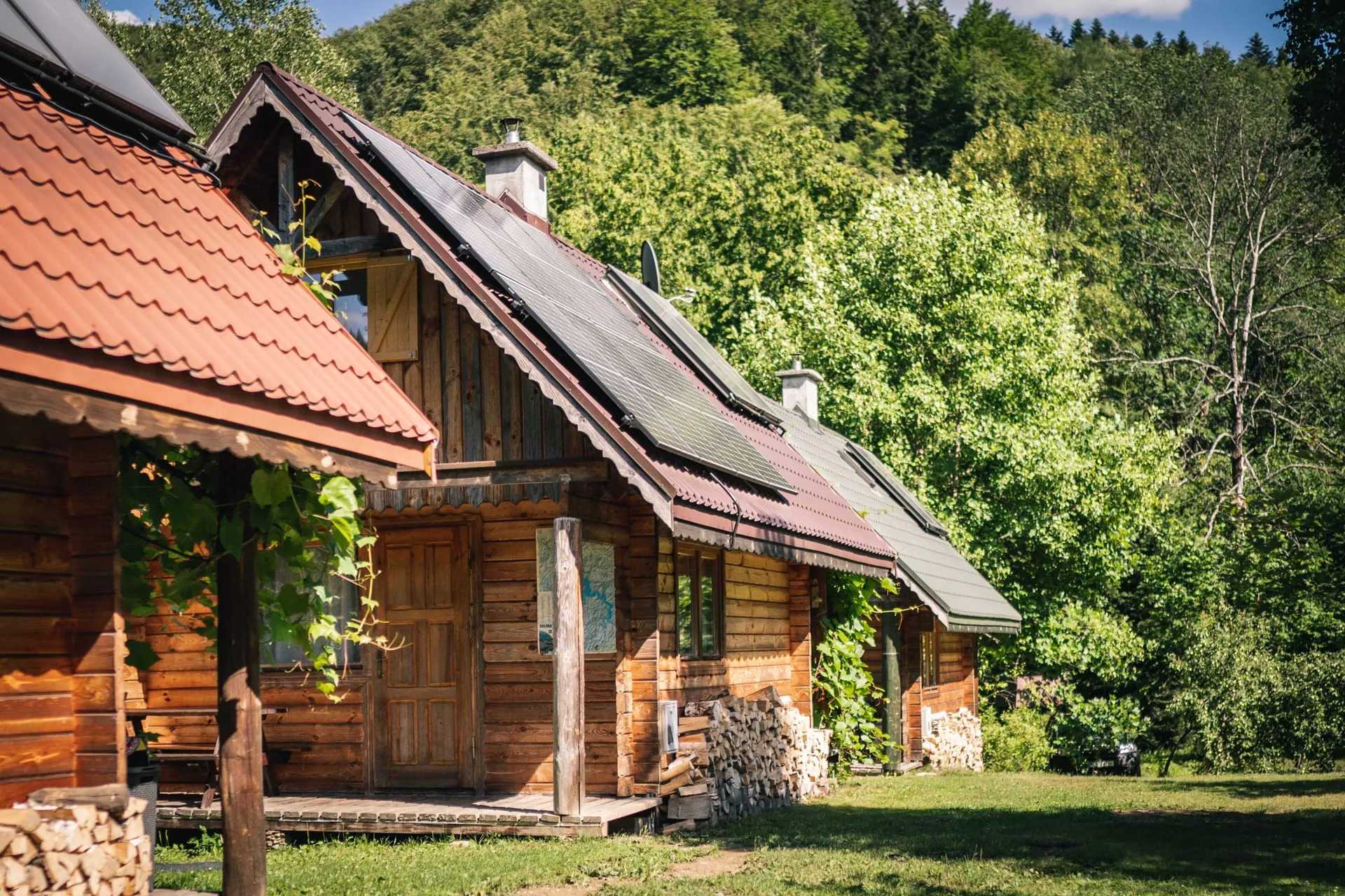 Rząd trzech drewnianych domków na słonecznej łące, za nimi zalesione wzgórze